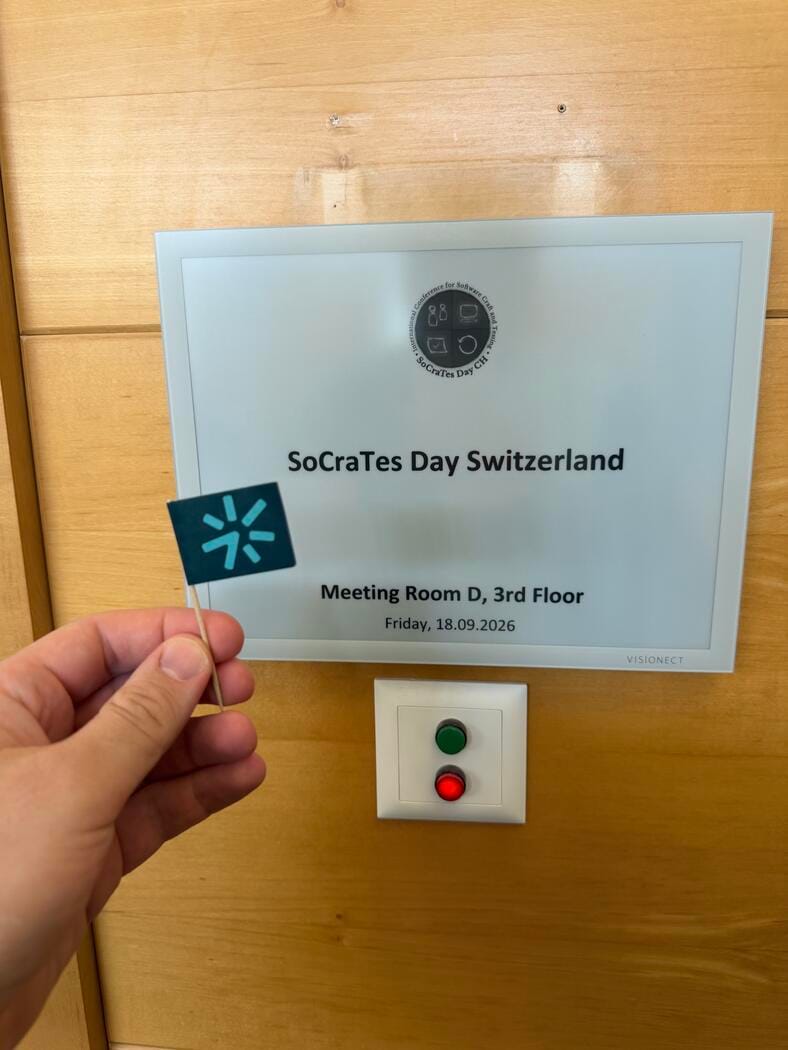 A door sign reading SoCraTes Day Switzerland, Meeting Room D, 3rd Floor, Friday, 18.09.2026, with the event logo. A hand in front holds a small paper flag with the easylearn logo.
